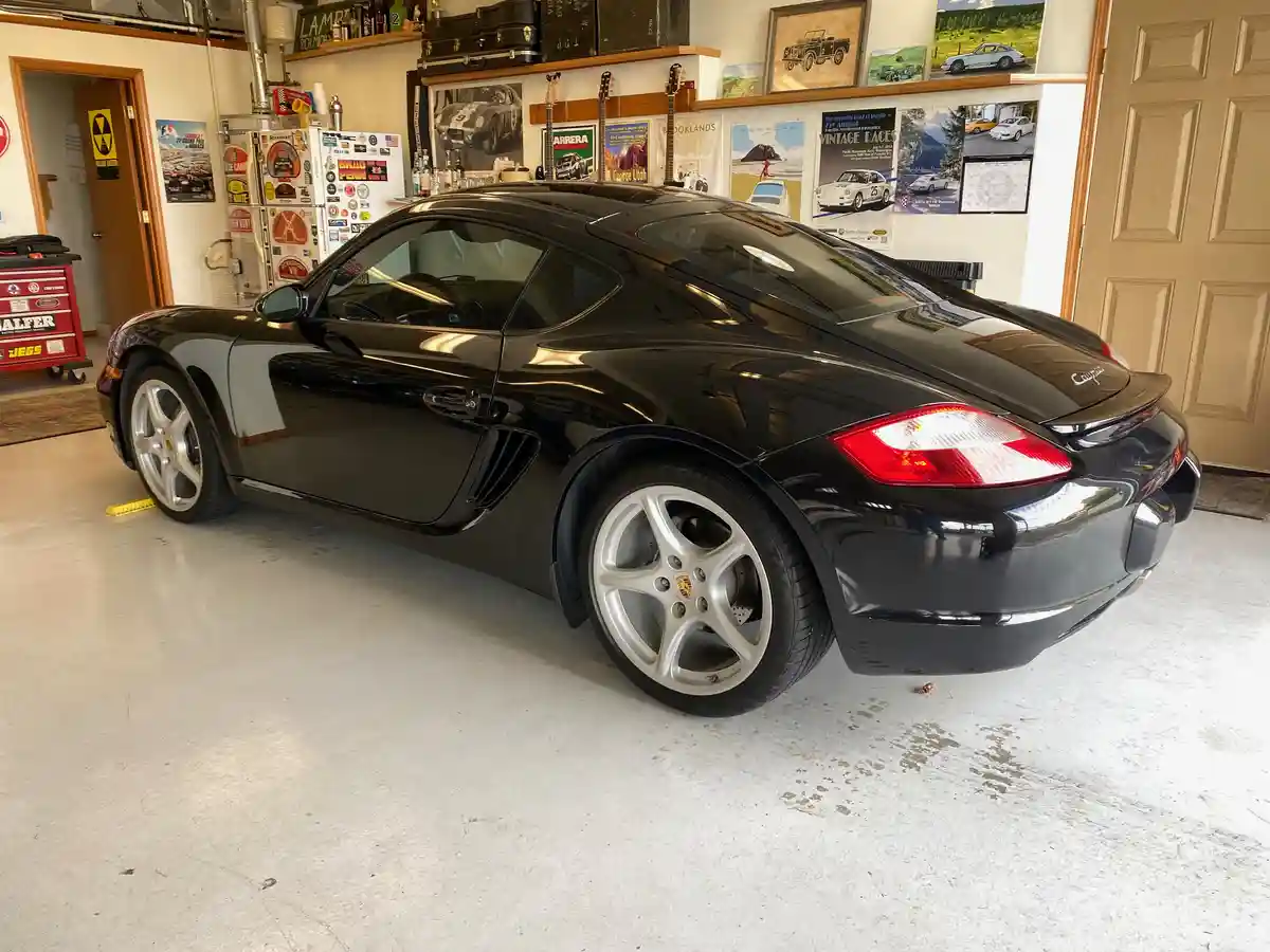 A black Porsche Boxster S is parked in a garage filled with automotive memorabilia, including posters, guitars, and a…