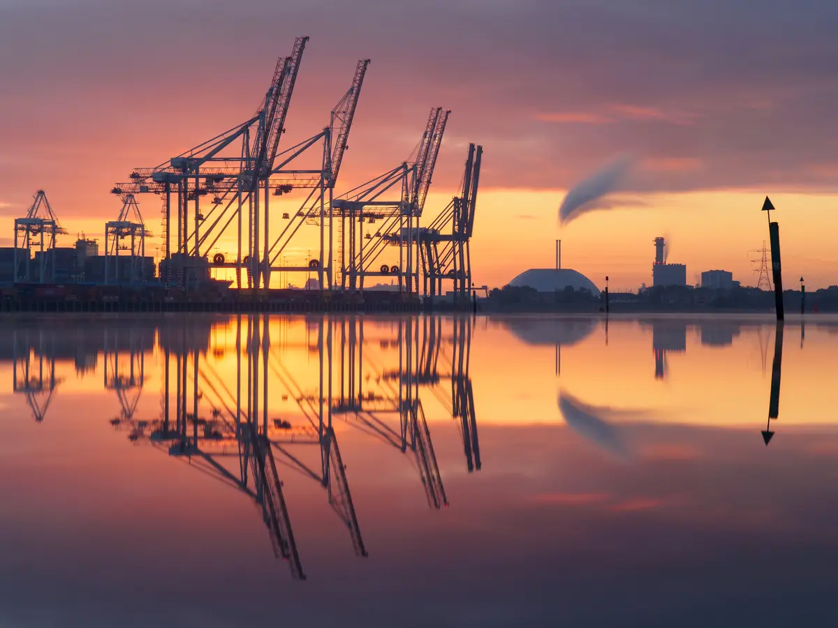 A serene sunset photograph shows large industrial port cranes silhouetted against a vibrant orange and purple sky, with…