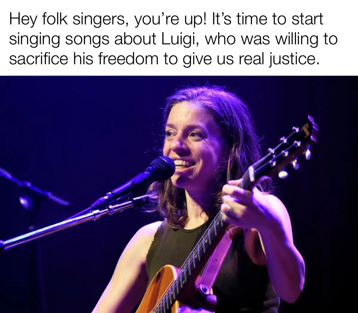 A woman with long brown hair, wearing a dark green sleeveless top, smiles while playing an acoustic guitar and singing into…