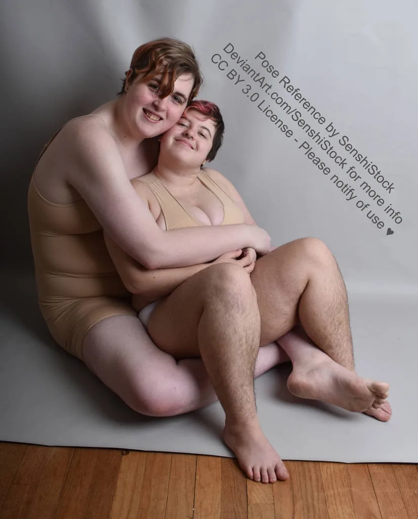 Two people are sitting on a gray floor mat against a plain gray backdrop, embracing each other affectionately. The person on…