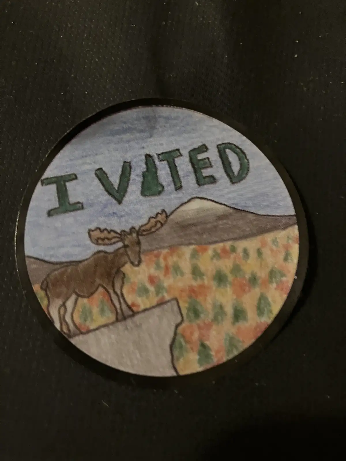 A hand-drawn circular badge depicts a moose standing on a rocky outcrop in a forested, mountainous landscape. The word 'I…