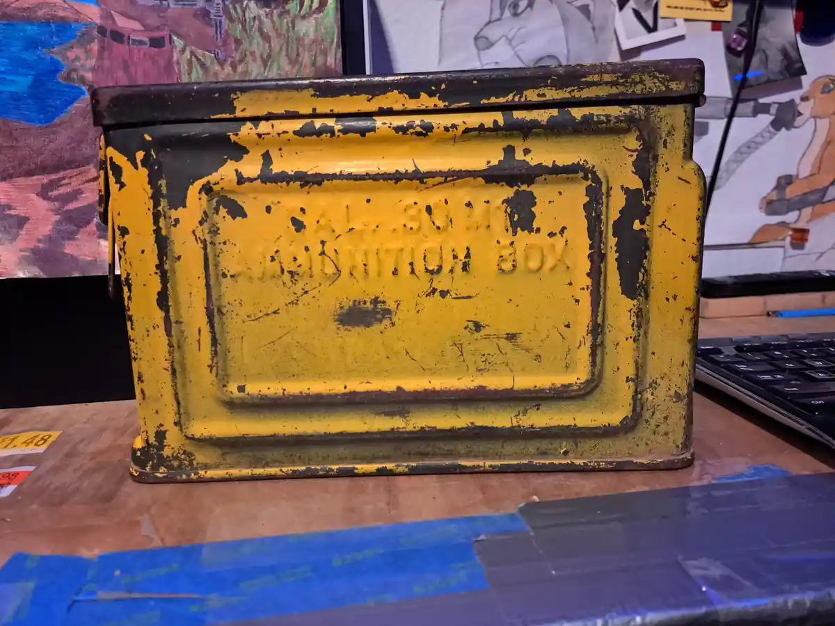 A weathered, yellow metal box with black paint peeling off, showing embossed text that reads 'CAL .30-06 AMMUNITION BOX'. It…