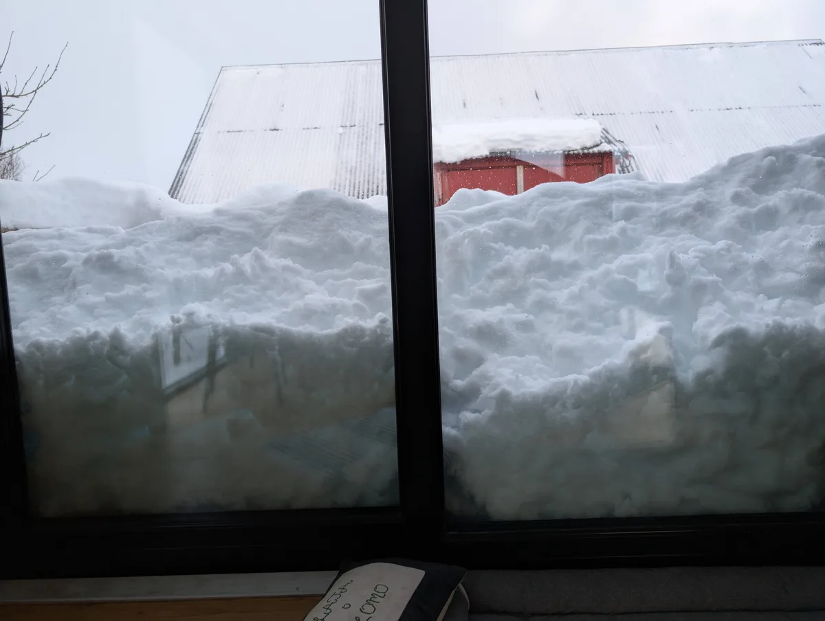 A view from inside a house looking out a large window at a massive, fluffy snowdrift that almost completely covers the view…