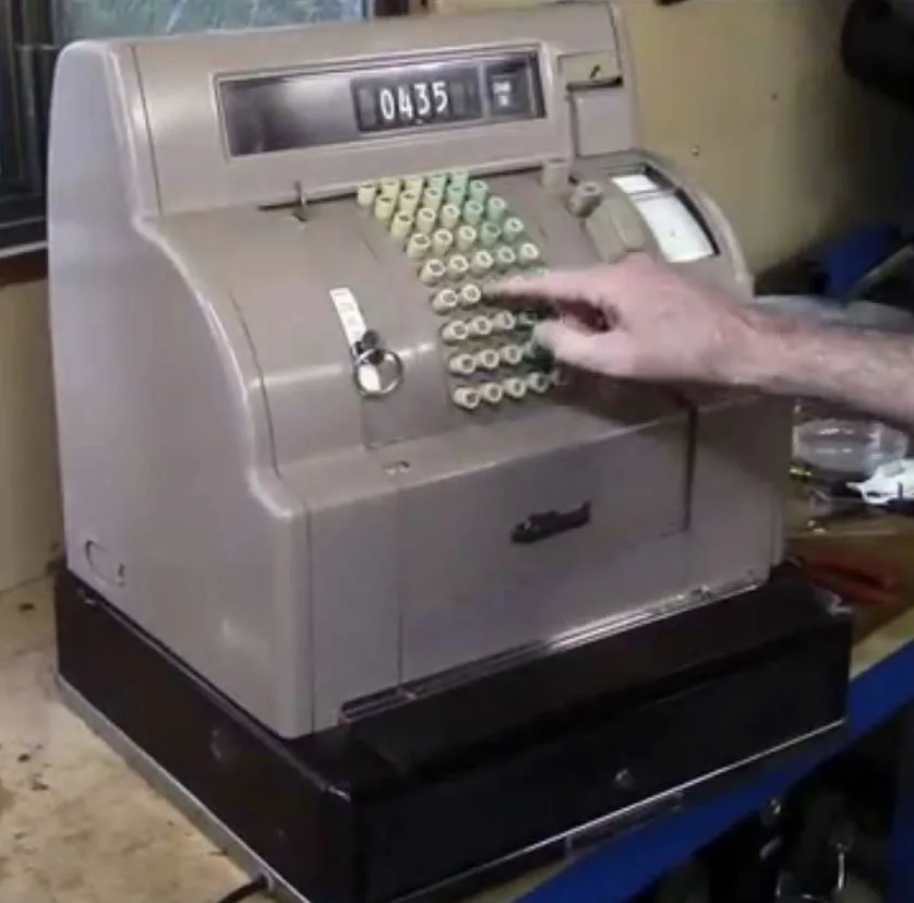 A person's hand presses a button on an old-fashioned beige mechanical cash register with a digital display showing '0435'…