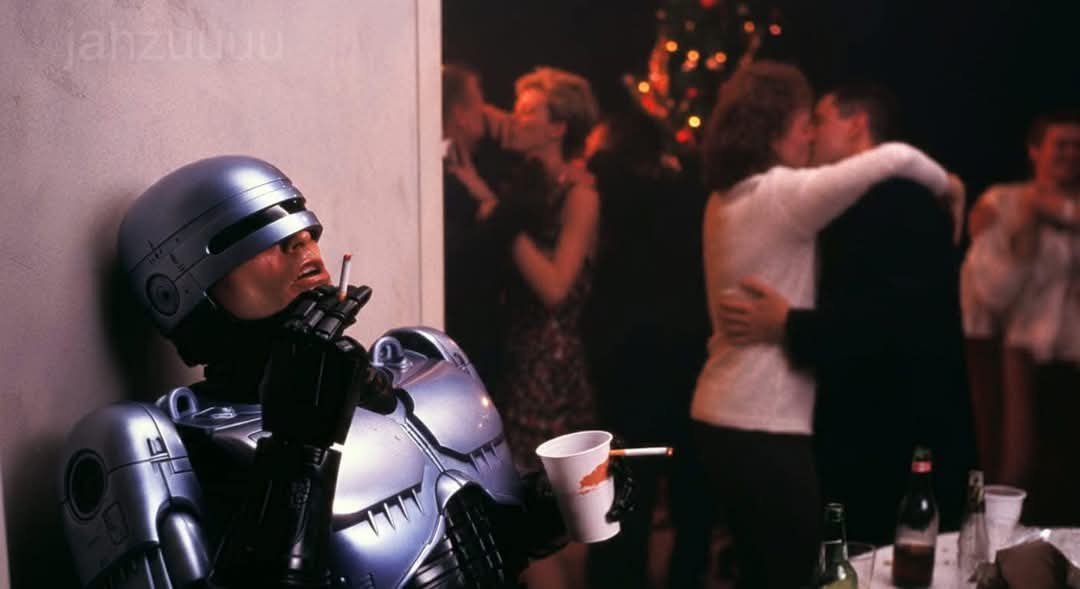 A silver robot from the movie RoboCop leans against a wall at a party, holding a cigarette and a paper cup, while blurred…