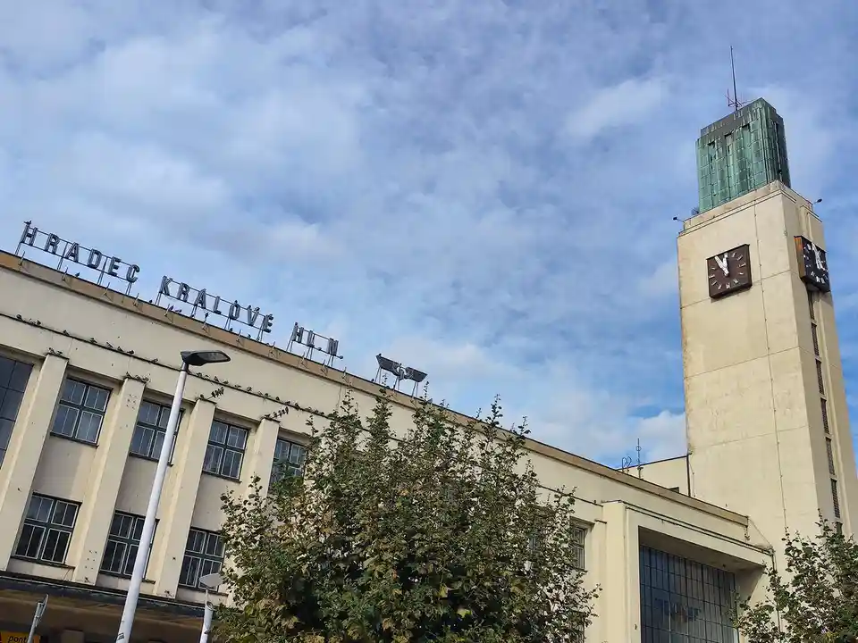 A low-angle photo of the Hradec Králové train station building under a partly cloudy sky, featuring its clock tower and…