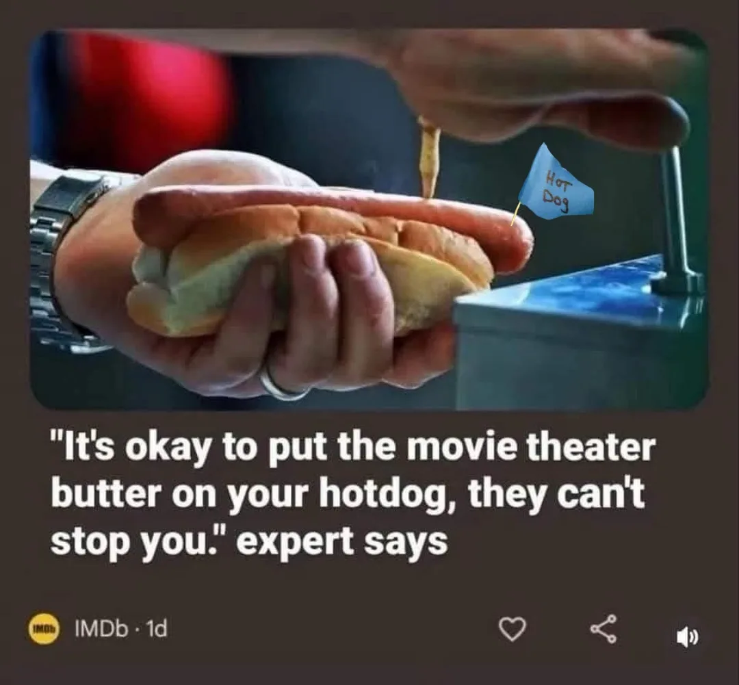 A person holds a hot dog in a bun while a stream of yellow butter is being squeezed onto it from a dispenser. A small blue…