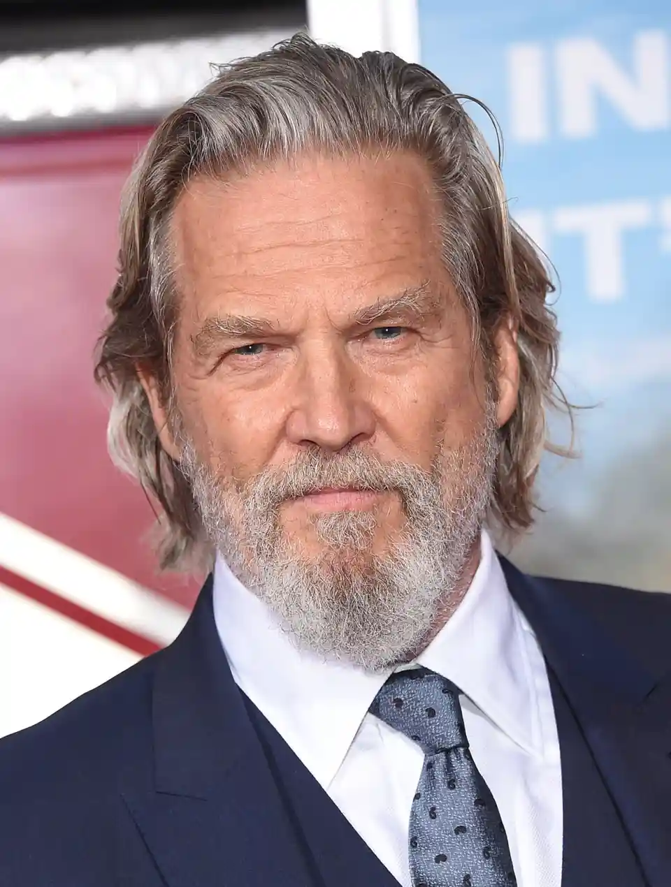 a close-up photo of actor Jeff Bridges at a red carpet event, wearing a dark suit, white shirt, and patterned tie, with his…