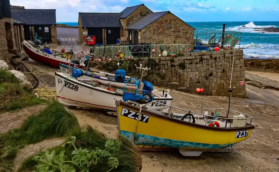 Three fishing boats are parked on a muddy shore next to stone buildings and a sea wall, with the ocean and waves visible in…