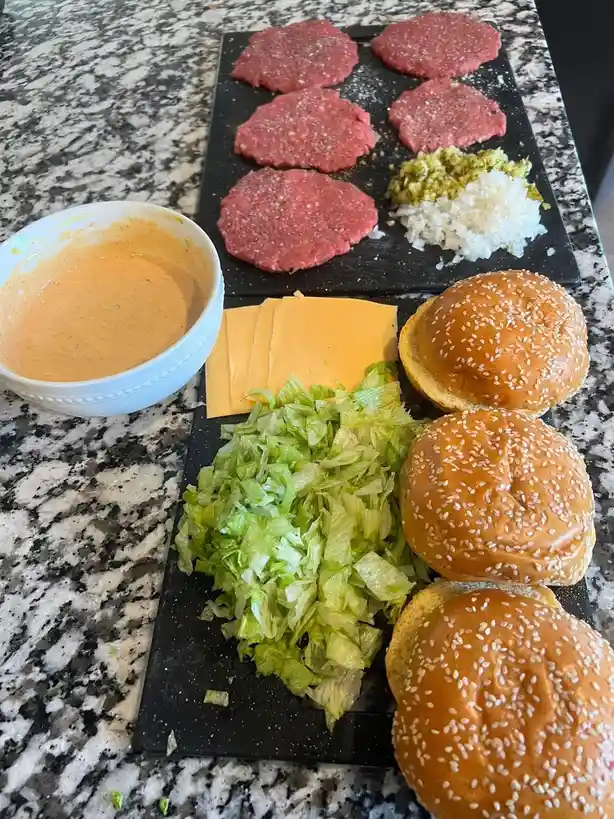 A kitchen counter displays ingredients for making burgers: six seasoned raw beef patties, chopped lettuce, cheese slices…