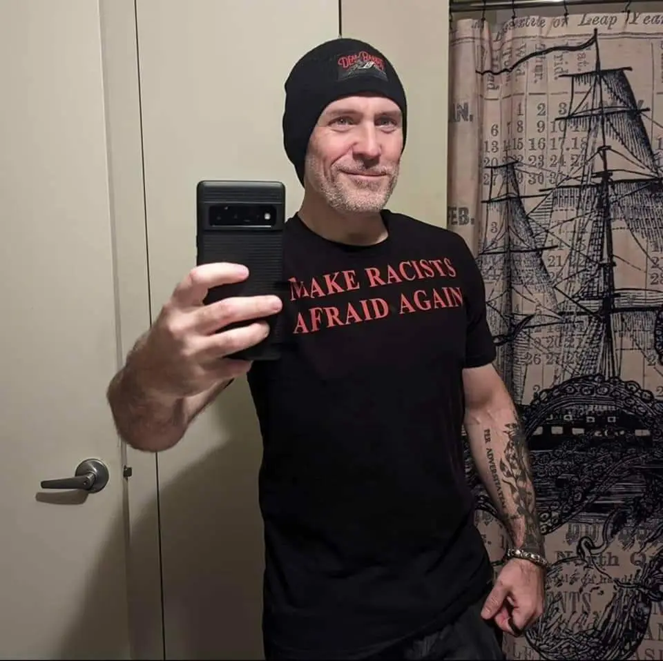 A man with a beard and black beanie takes a mirror selfie holding a black smartphone, wearing a black t-shirt with red text…