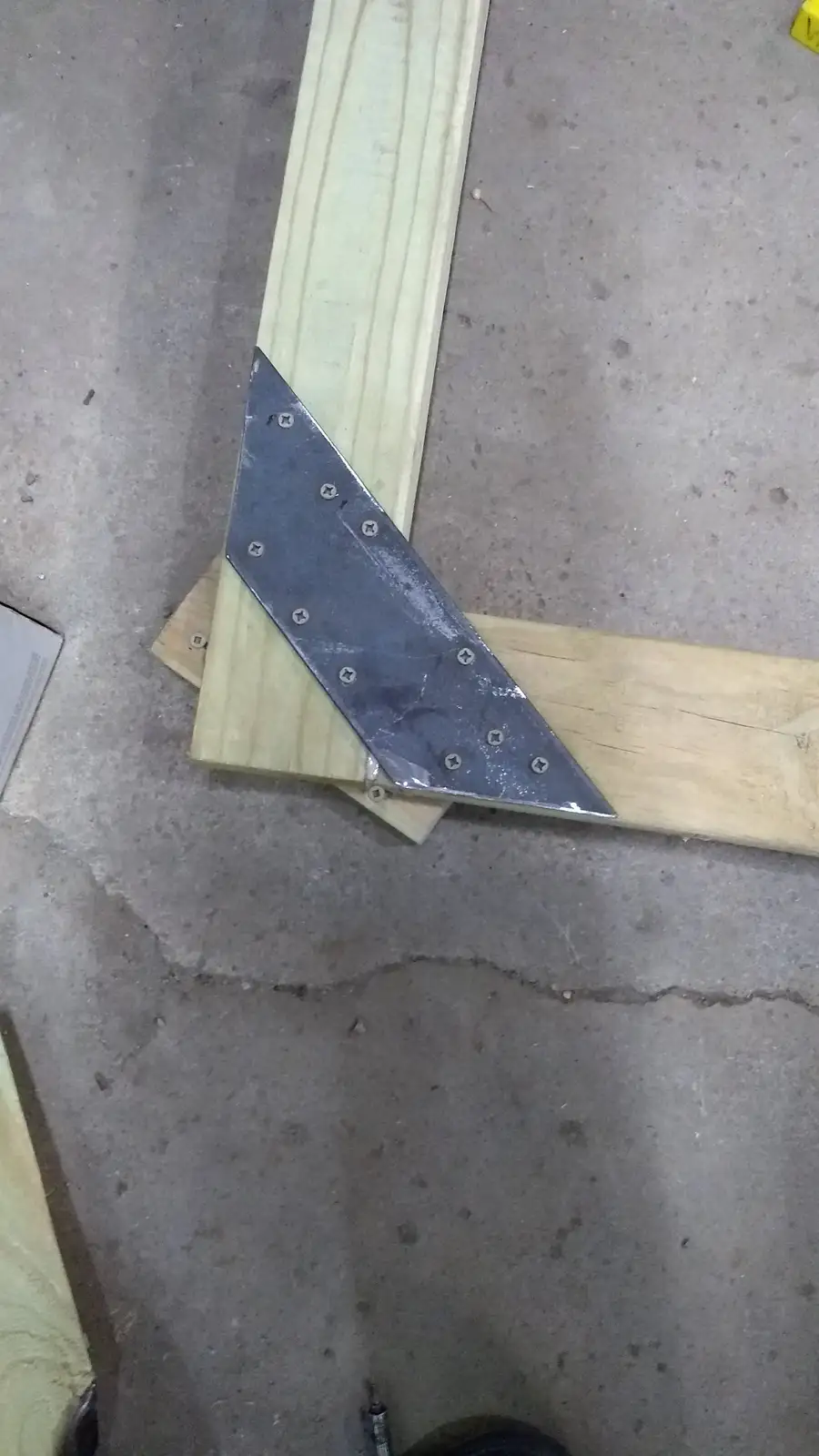 A metal triangular ruler, bolted with multiple screws, is placed on two wooden planks forming a right angle on a concrete…