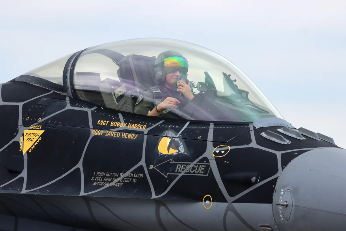 A close-up of a fighter jet's cockpit shows a smiling pilot in flight gear and a reflective helmet. The jet's fuselage is…