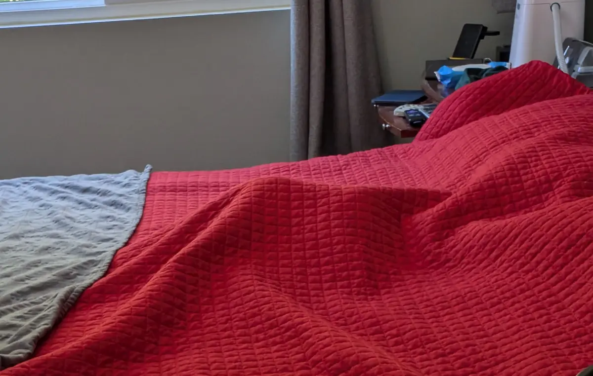 A close-up photo of a rumpled red quilted bedspread with a gray blanket partially folded over it, in a bedroom with a…