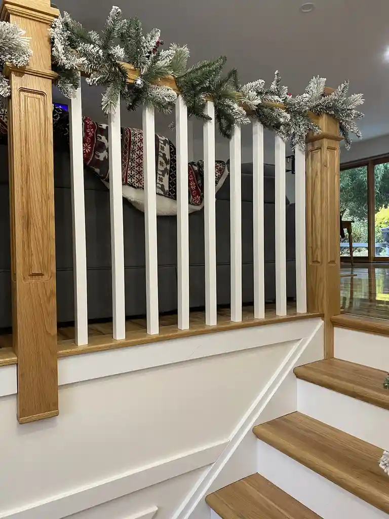 A staircase with a wooden railing decorated with a flocked green garland and silver ornaments. Behind the railing, a dark…
