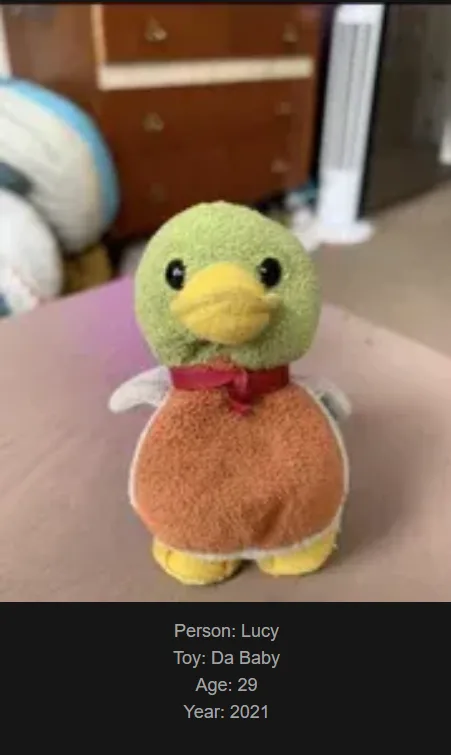 A green towel duck plush toy wearing an orange towel dress and a red ribbon around its neck, sitting on a pink surface. Text…