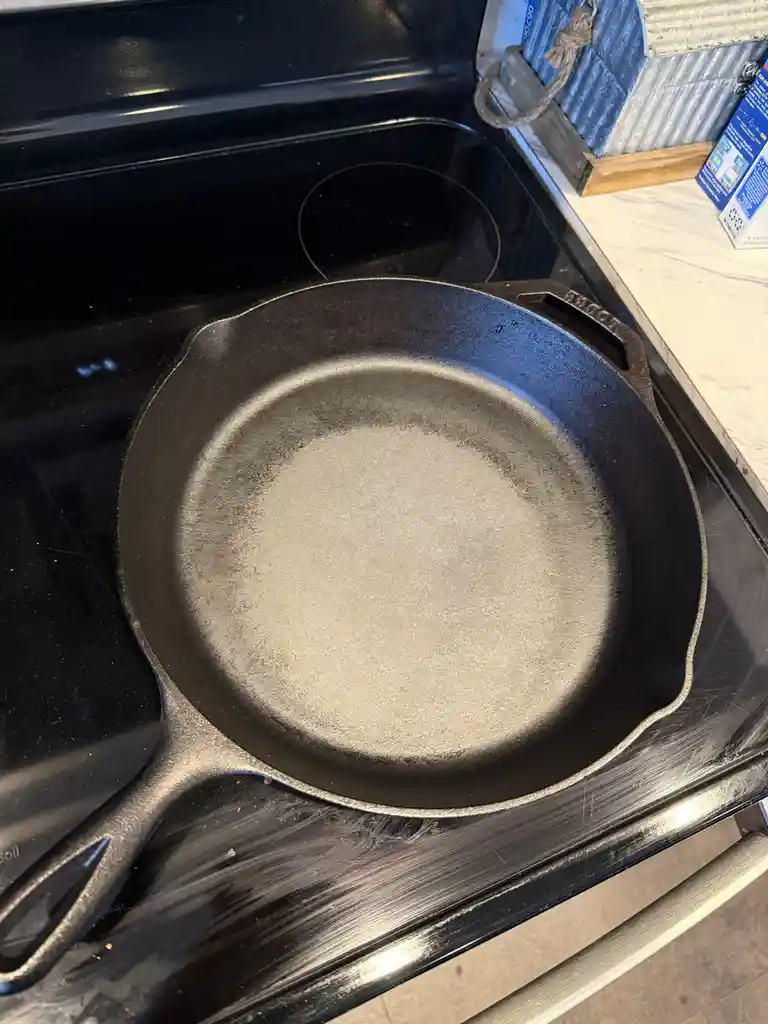 A black cast iron skillet sits on a black electric stovetop, its cooking surface covered in a thick, light-gray powdery…