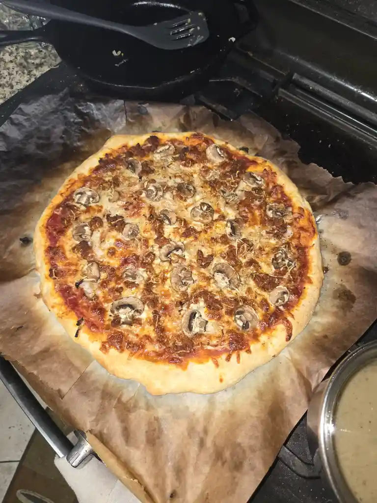 A homemade pizza with mushrooms and cheese sits on parchment paper on a stovetop, with a cast iron skillet and spatula…