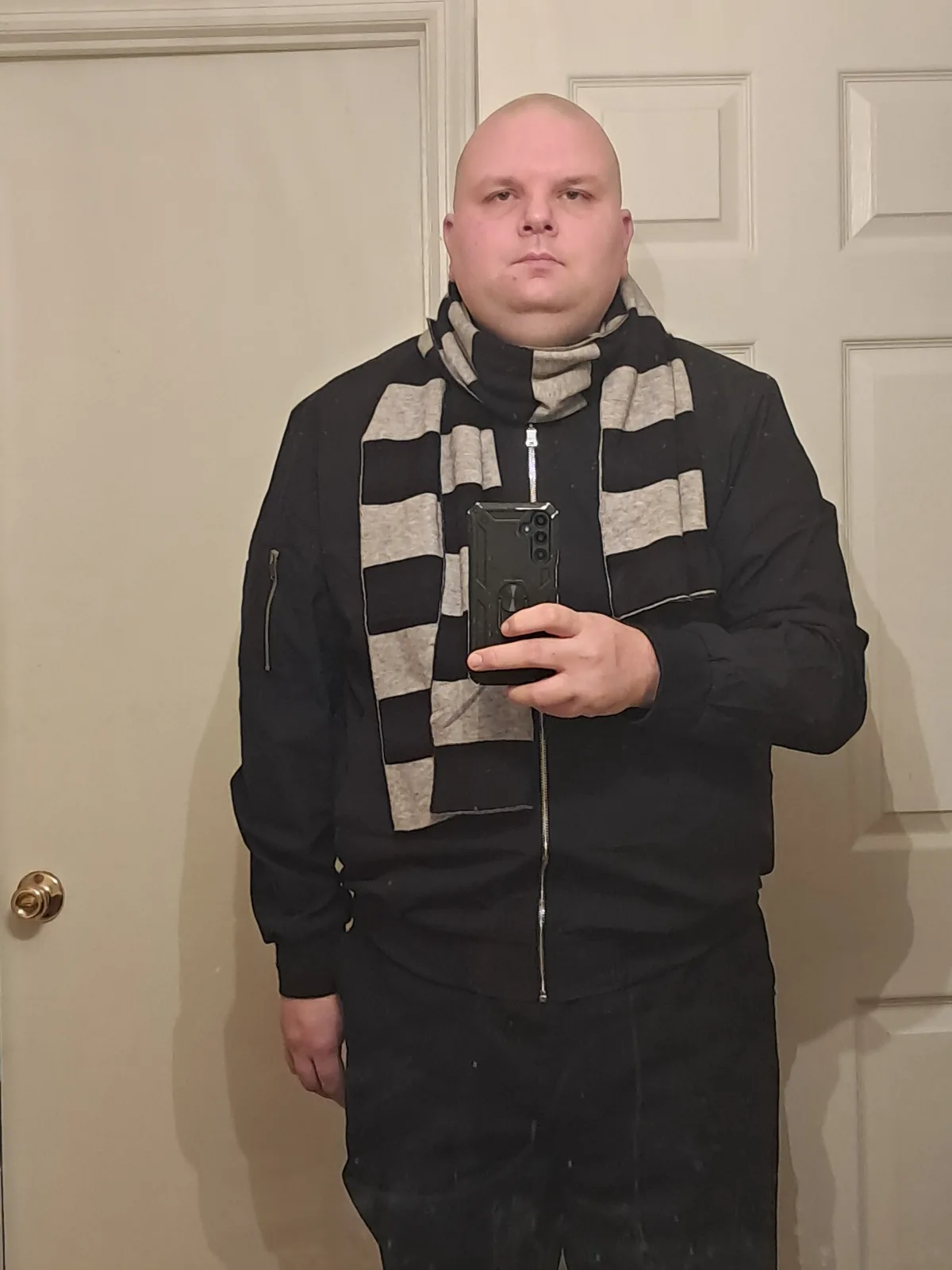 A bald man in a black zip-up jacket and black pants stands in front of a white door, holding a phone to take a selfie. He…