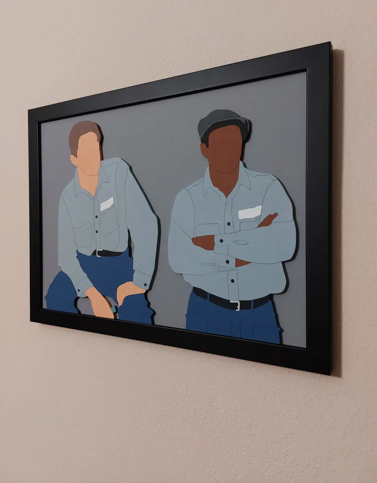 A framed, stylized artwork on a wall depicts two men in light blue work shirts and blue pants, both with blank faces. The…