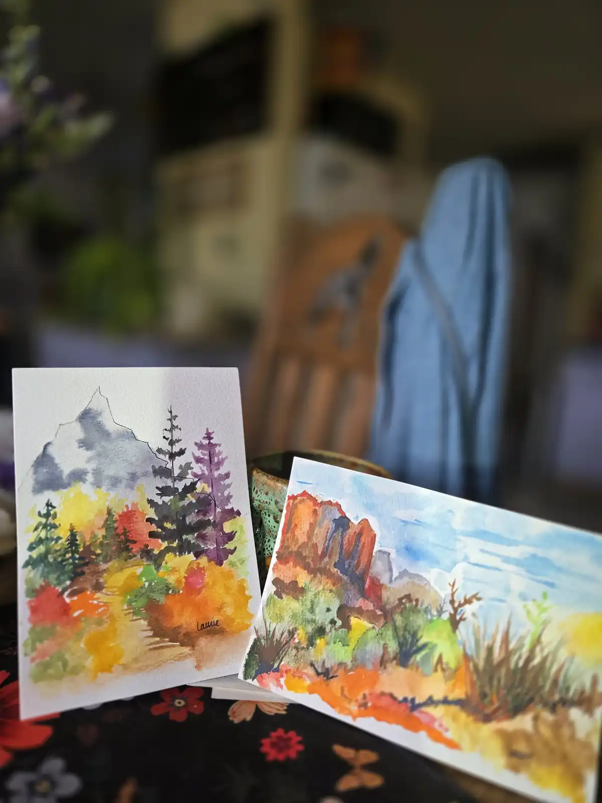 Two watercolor landscape drawings are propped up on a floral tablecloth, one depicting snowy mountains with colorful trees…