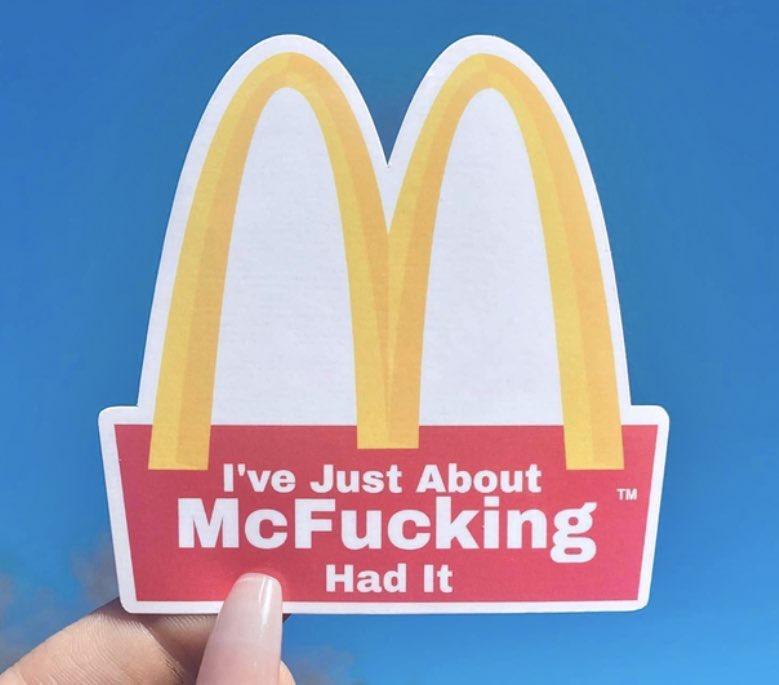 A hand holds a cutout of the McDonald's golden arches logo against a blue sky. The red base of the logo has white text that…