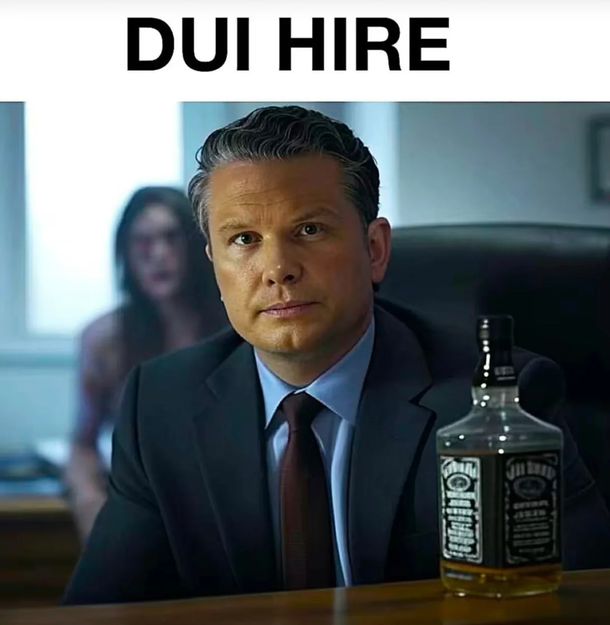 A man in a suit and tie sits at a desk with a bottle of Jack Daniels whiskey in front of him, looking serious. Behind him, a…