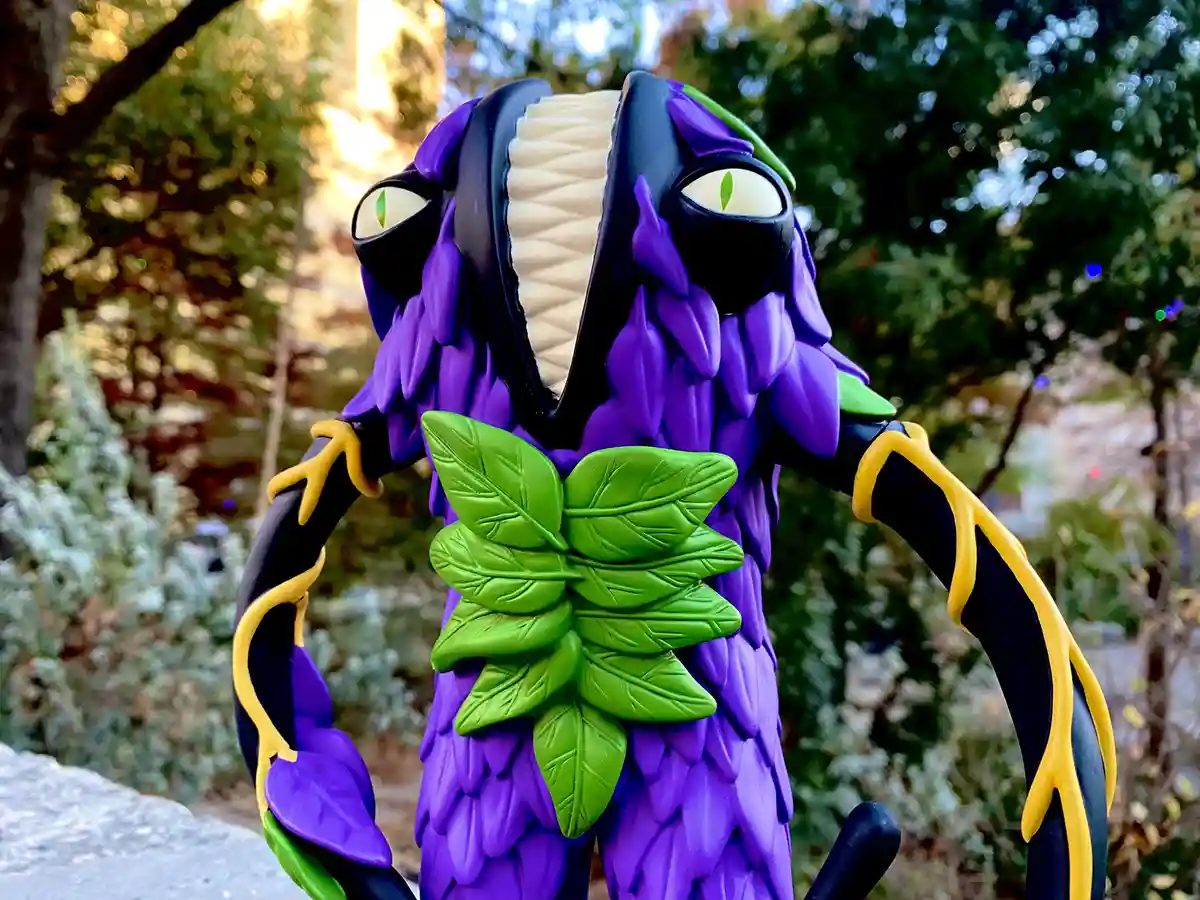 A purple, leafy monster figurine with green eyes and a wide, toothy grin stands outdoors with trees in the background. It…