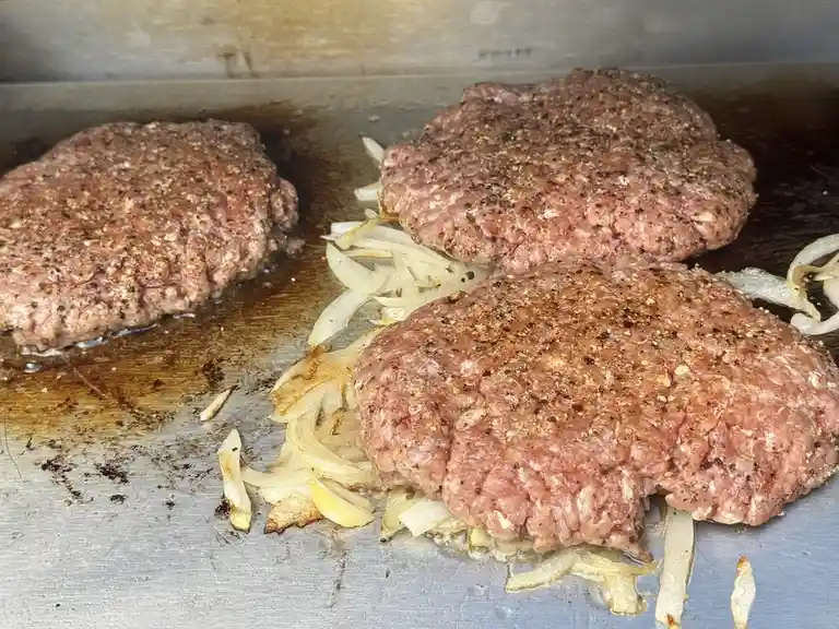 Three raw, seasoned hamburger patties are cooking on a metal griddle alongside caramelized onions. The text overlay reads…