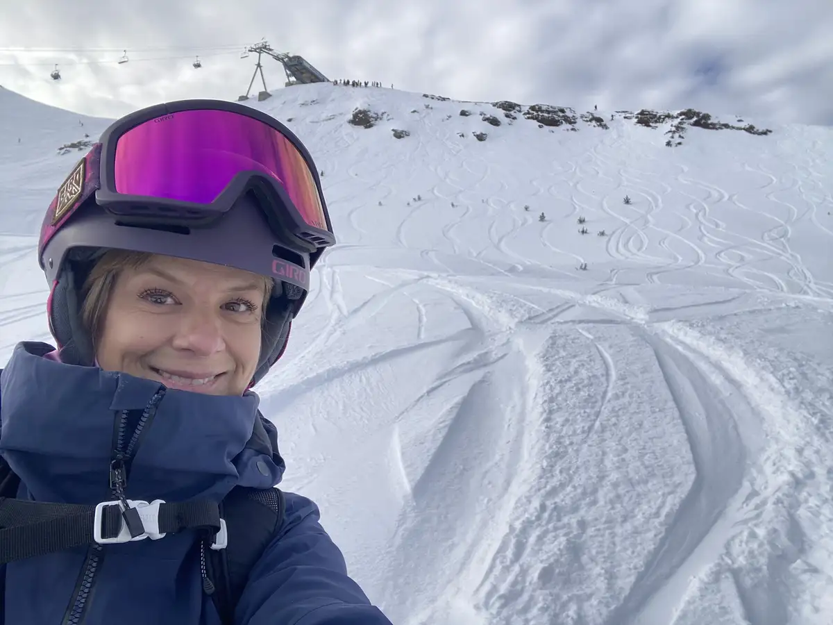 A woman wearing a ski helmet and goggles smiles at the camera while standing on a snowy mountain slope covered in numerous…