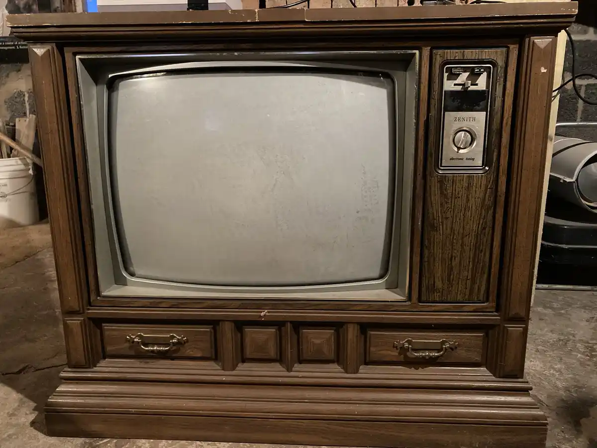 A vintage Zenith television with a wooden cabinet and silver control panel, its screen is blank and dusty, sitting in what…