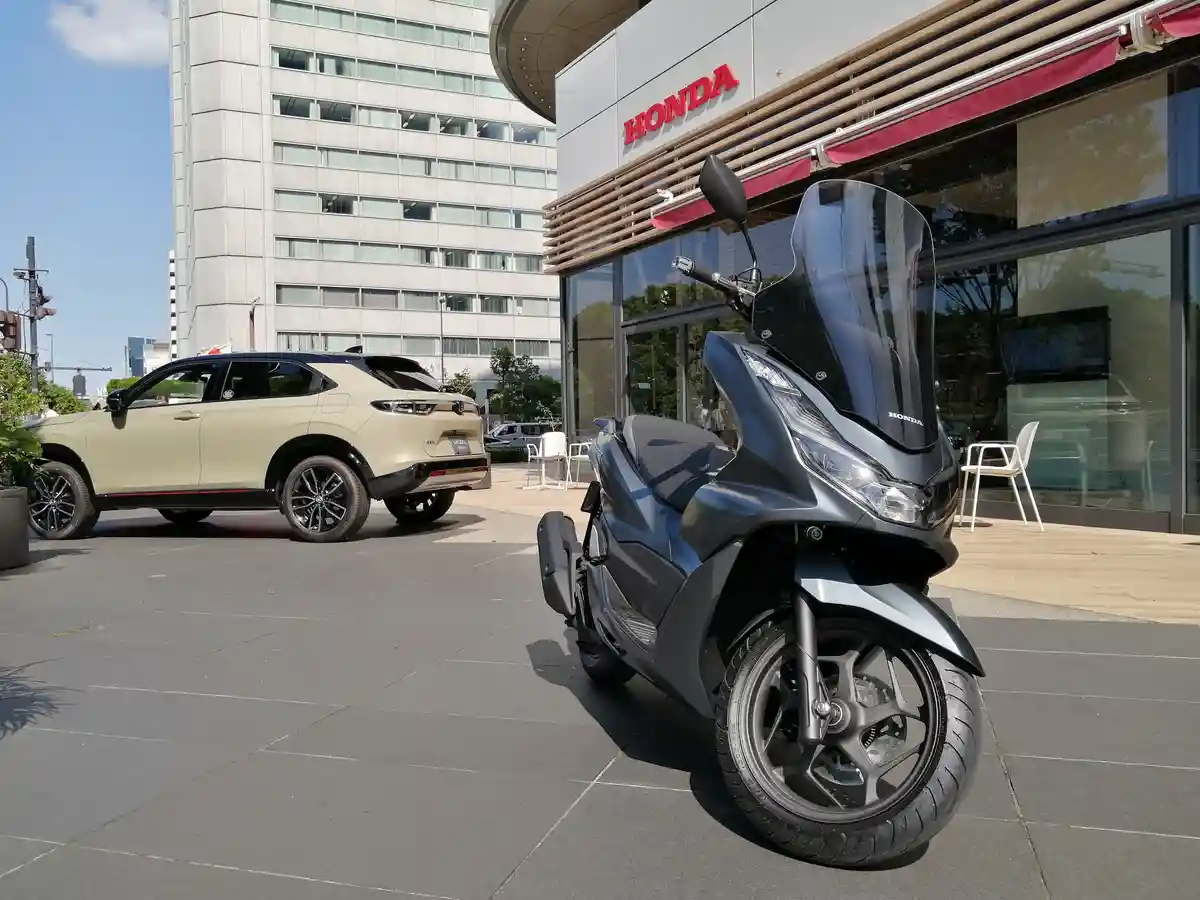 A beige Honda Vezel SUV is parked next to a dark gray Honda scooter in front of a Honda dealership. The scooter is in the…