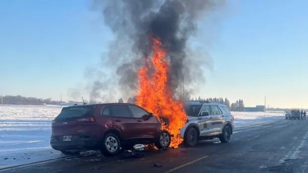 Two cars are severely damaged and engulfed in flames on a snowy road, with thick black smoke rising into the clear blue sky…