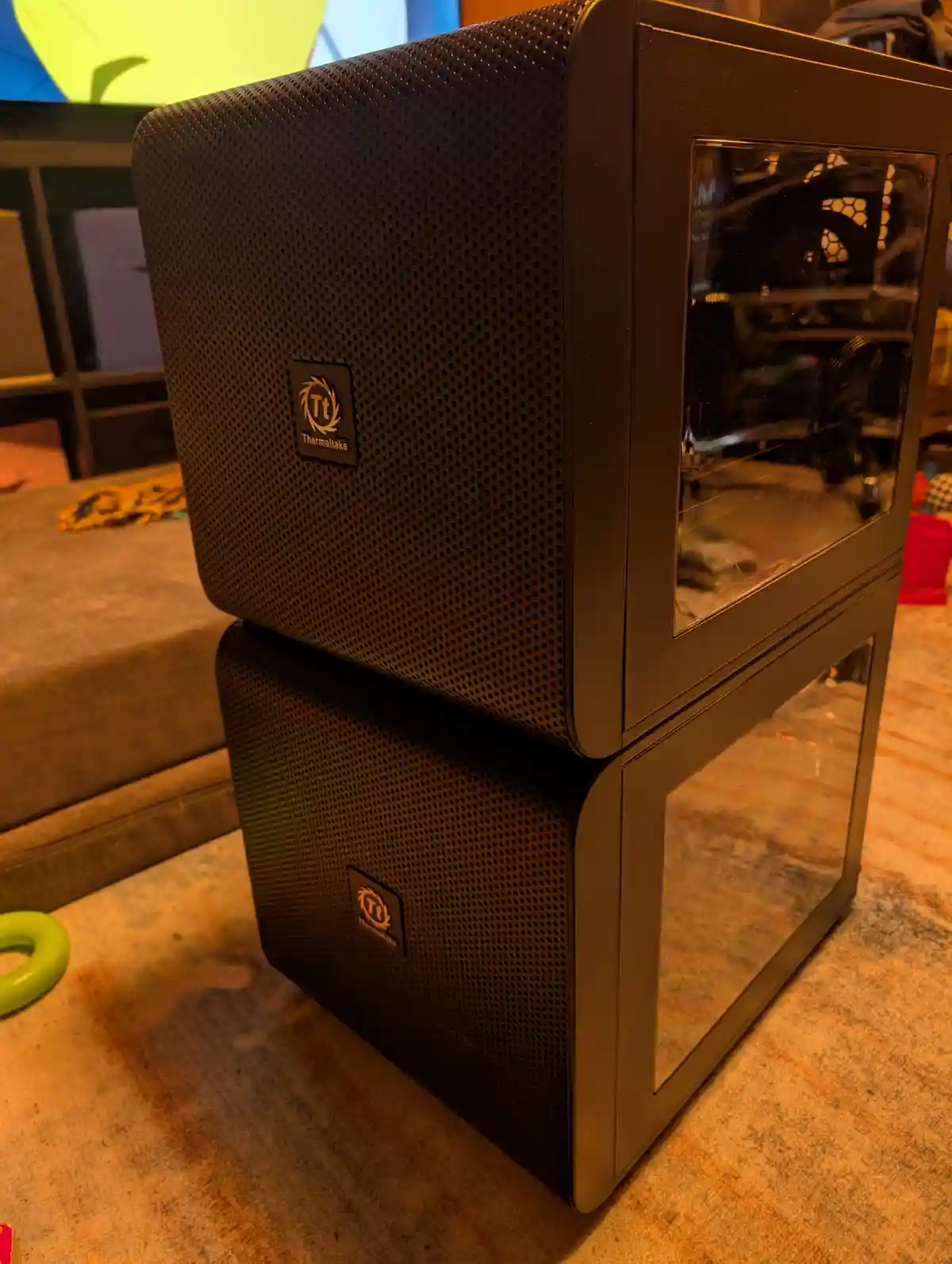 Two black Thermaltake computer cases are stacked vertically on a carpeted floor, with the top case resting directly on the…