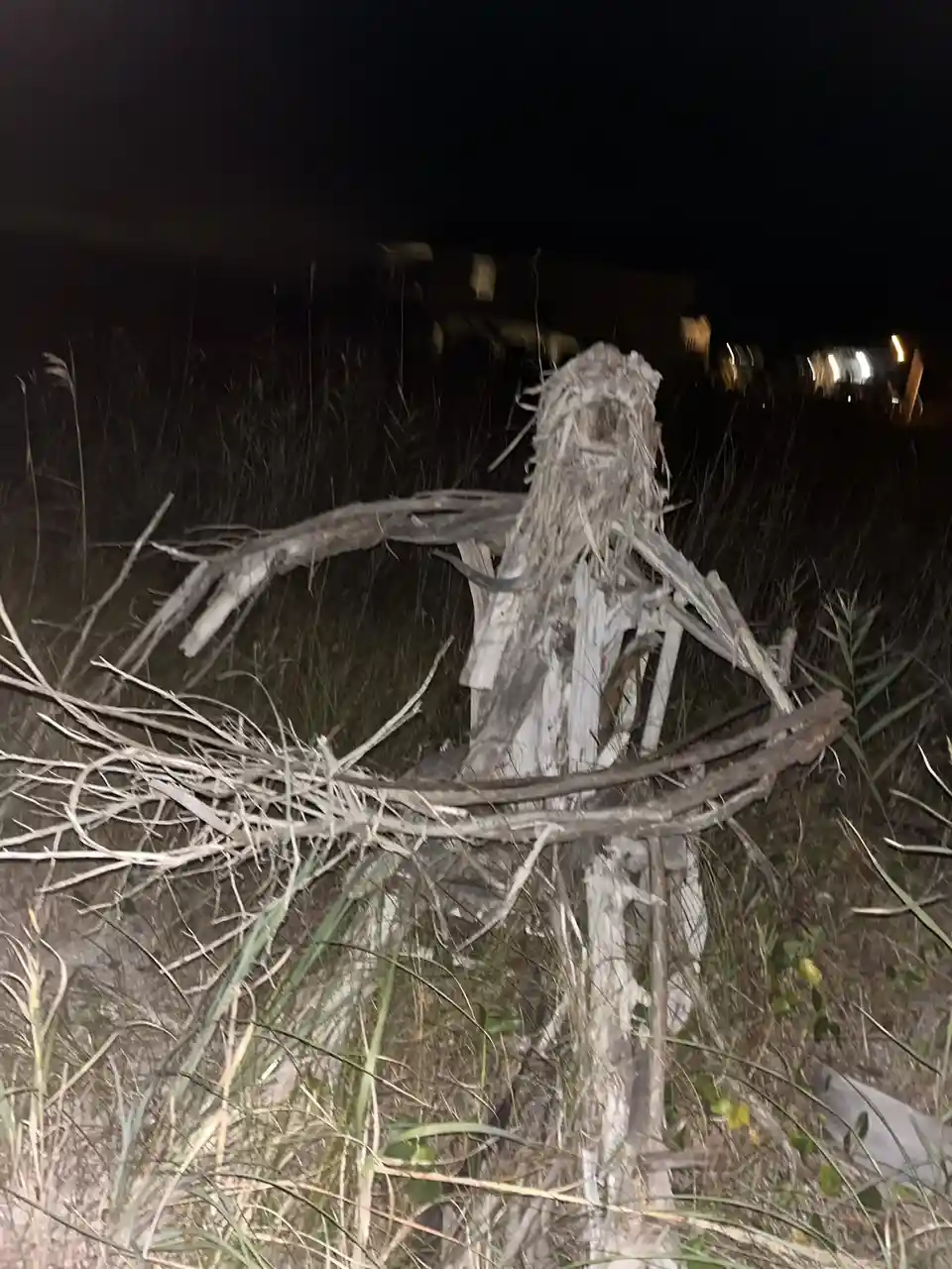 A dilapidated scarecrow made of sticks and dried grass stands in a field at night, its limbs askew and head askew, with a…