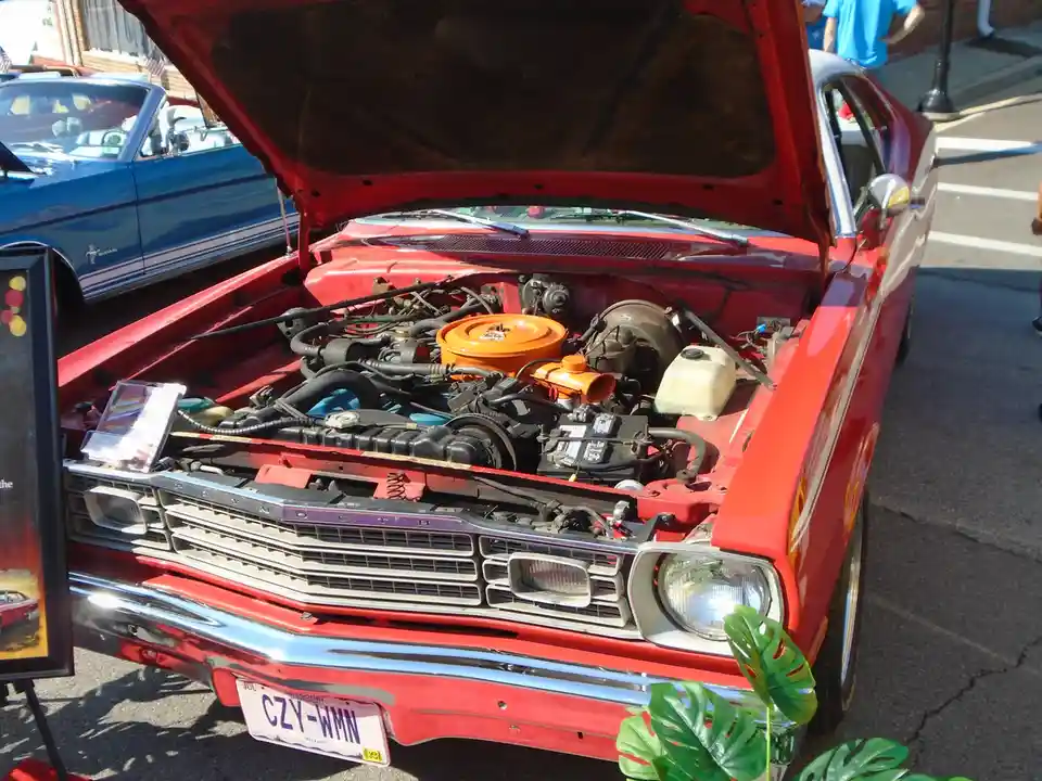 A red classic Plymouth car with its hood open, revealing a detailed engine with an orange air filter, parked outdoors at…