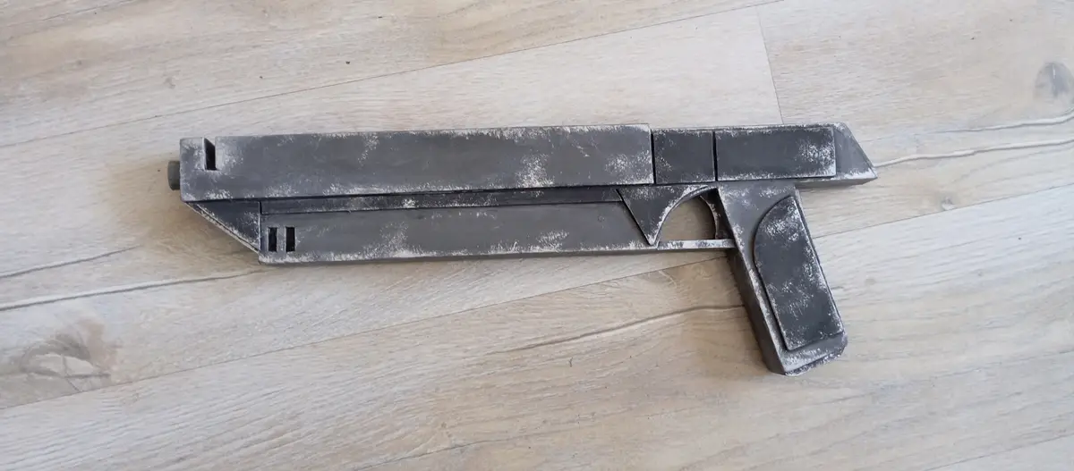 A crudely constructed, blocky gun prop made from what appears to be wood and metal, resting on a light-colored wooden floor…