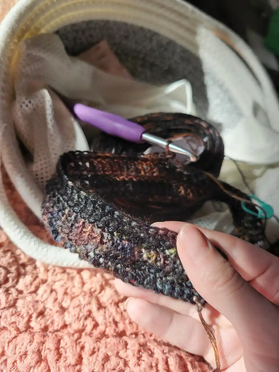 A hand holds a partially completed, multicolored crocheted item with a purple crochet hook resting in a basket filled with…