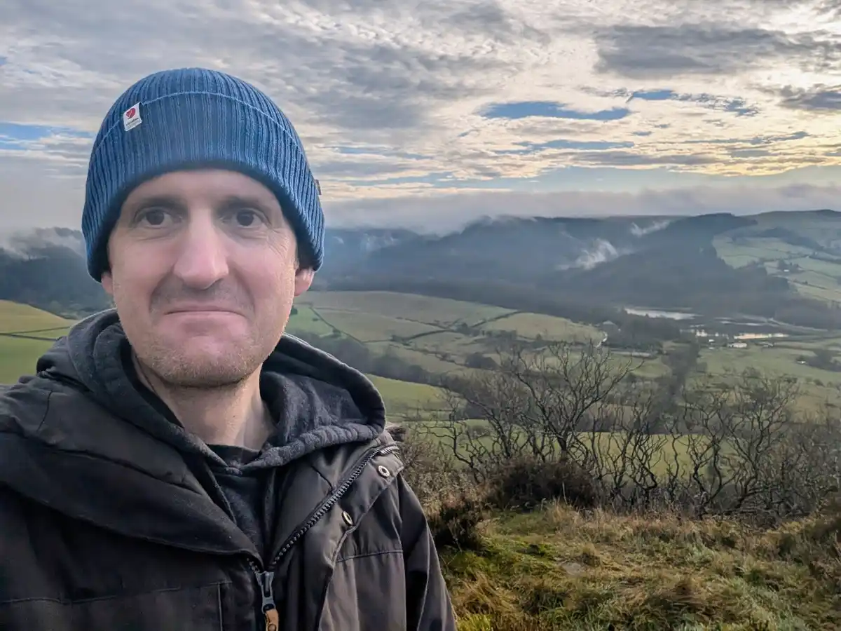A man wearing a blue beanie and dark jacket stands on a hillside overlooking a misty, rolling countryside with fields and…