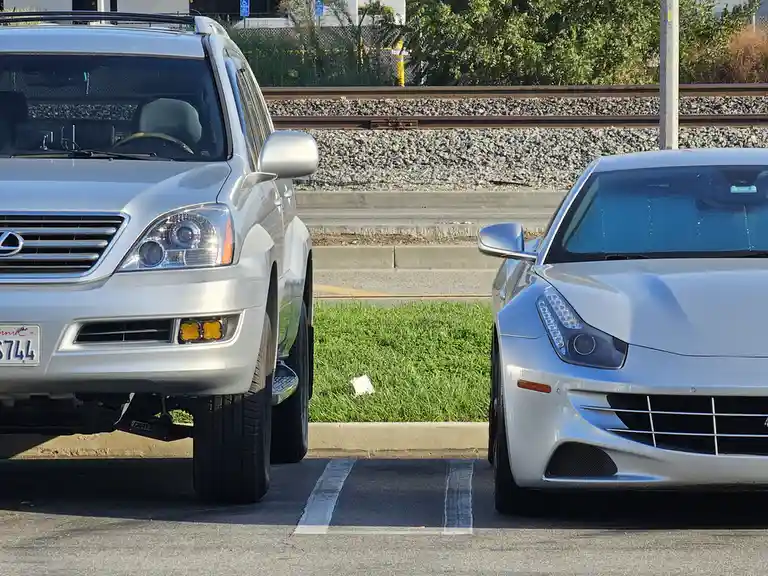 Two silver cars are parked side-by-side in a lot, with a silver Lexus SUV on the left and a silver Ferrari sports car on the…