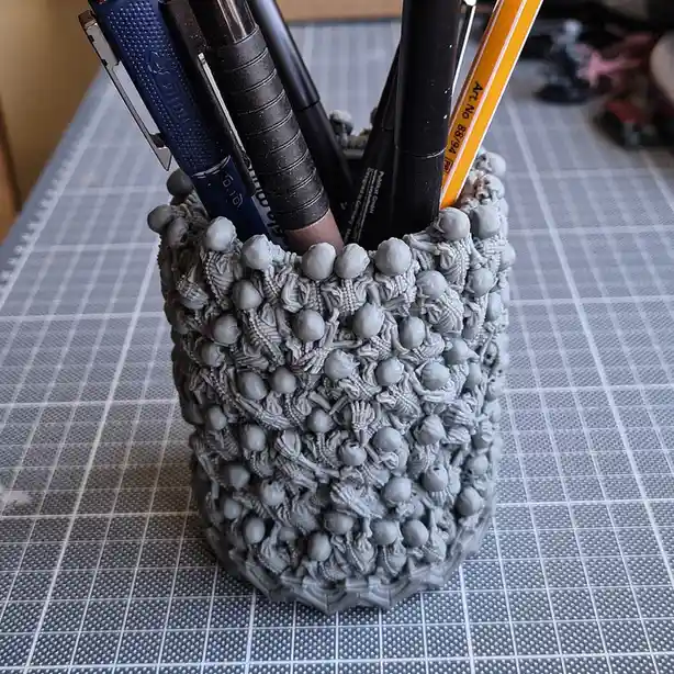 A gray, intricately textured pen holder made from 3d-printed miniature skeletons is filled with pens and pencils, sitting on…