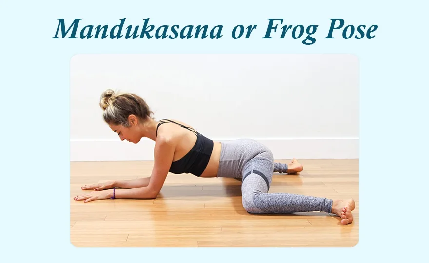 A woman in a black sports bra and grey leggings is on a wooden floor in a yoga pose with her knees bent wide and her torso…