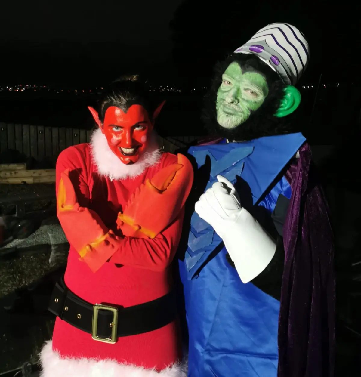 Two people in costumes pose at night. One is dressed as a red devil with horns and a Santa suit, arms crossed. The other is…