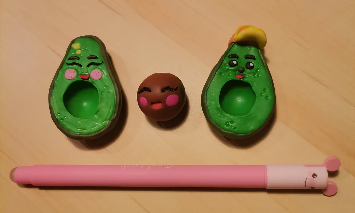 Three clay figurines on a wooden surface: two avocado halves with faces (one winking, one wearing a yellow hat) and a small…
