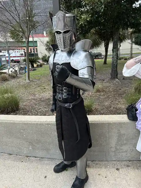 A person in full silver medieval knight armor, including helmet and gauntlets, stands outdoors in a park-like setting. They…