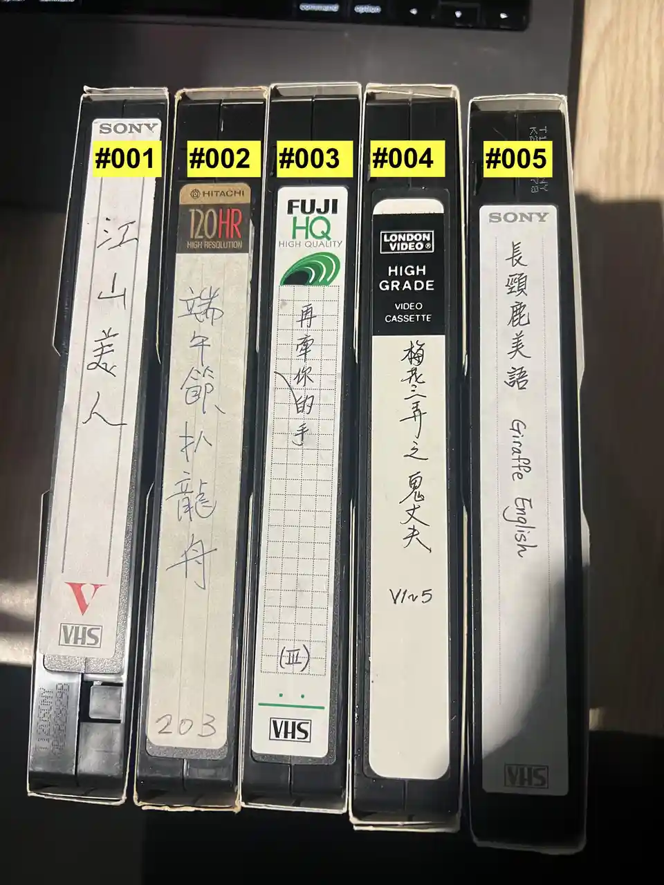 Five VHS tapes are lined up, each labeled with a yellow number sticker (#001 to #005) and handwritten Chinese characters on…