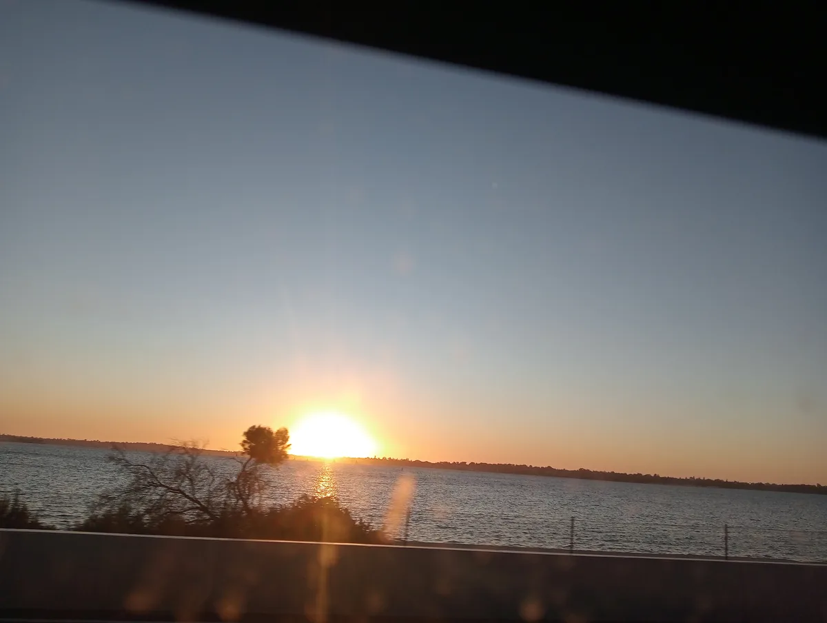 A scenic photo taken from inside a vehicle, showing a sunset over a large body of water with a distant shoreline. The sun is…