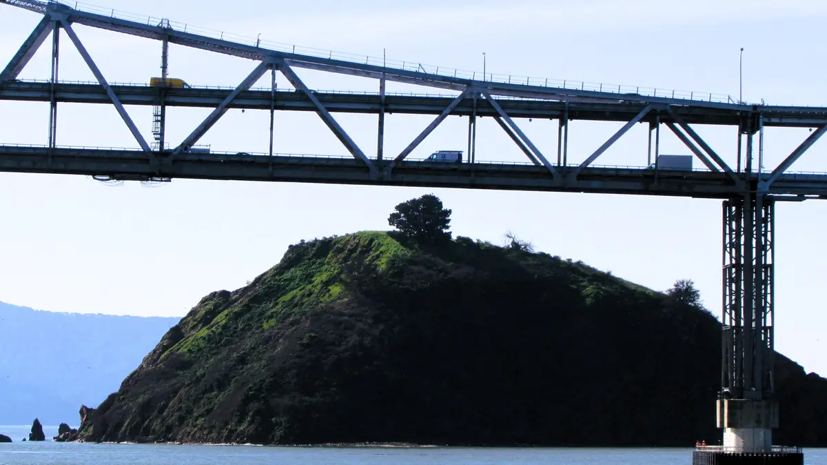 A large steel truss bridge spans across a body of water, with vehicles visible on its roadway. Below the bridge, a small…
