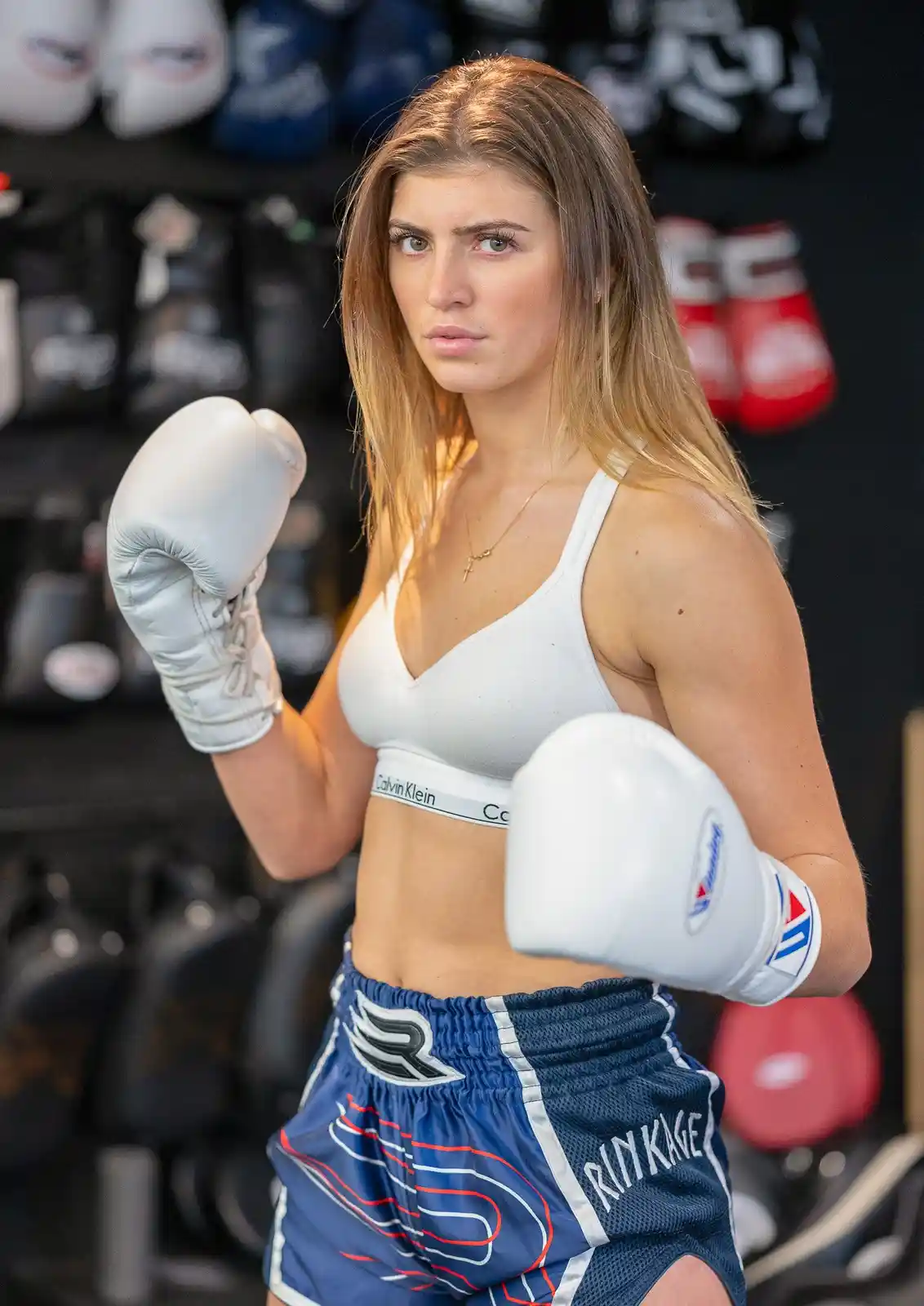 A woman in a boxing gym wearing white boxing gloves, a white Calvin Klein sports bra, and blue Rinkage shorts, strikes a…