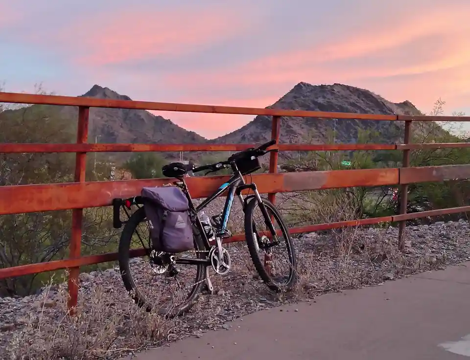 A mountain bike with a purple pannier bag leans against a rusty orange metal fence on a gravel path. Behind it, desert…